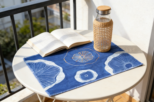 Open book on a table with a blue decorative mat, glass bottle, and outdoor setting.