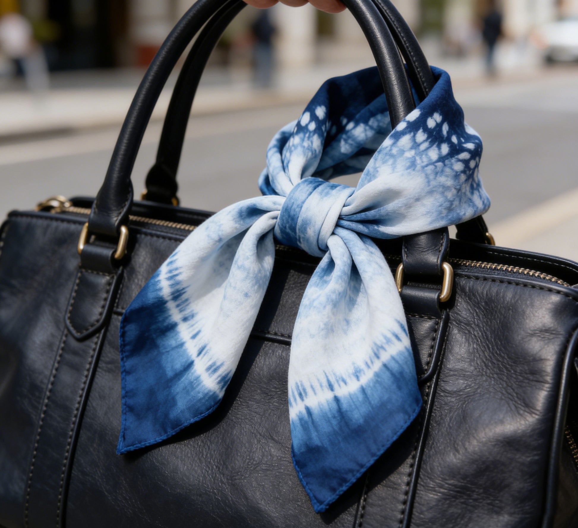 Black leather handbag with a blue and white tie-dye scarf tied around the handle, on a blurred street background.