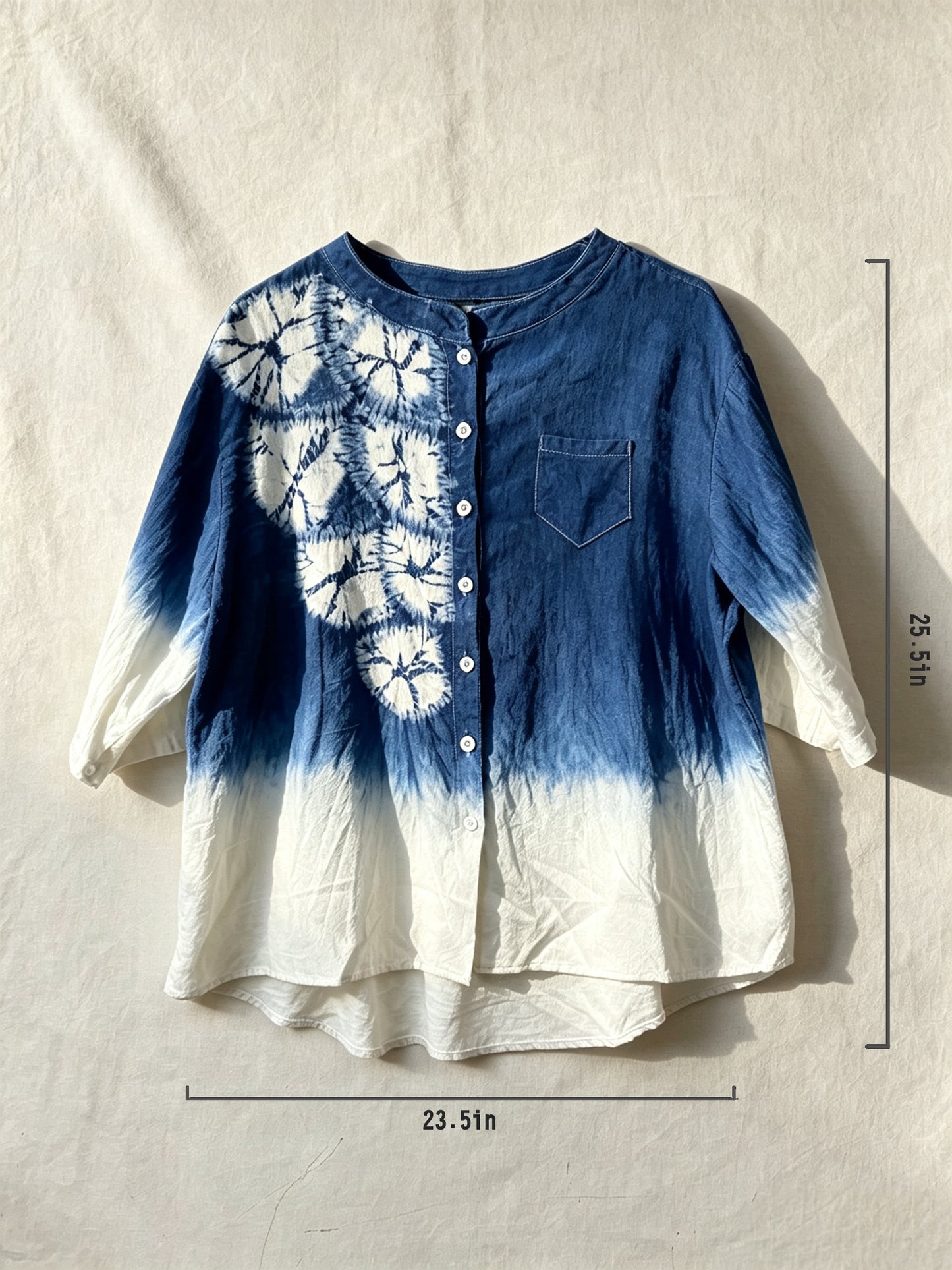 A blue and white tie-dye button-up shirt with a floral pattern on the upper left chest, laid flat on a textured beige fabric background with dimension lines indicating a height of 23.5 inches and width of 23.