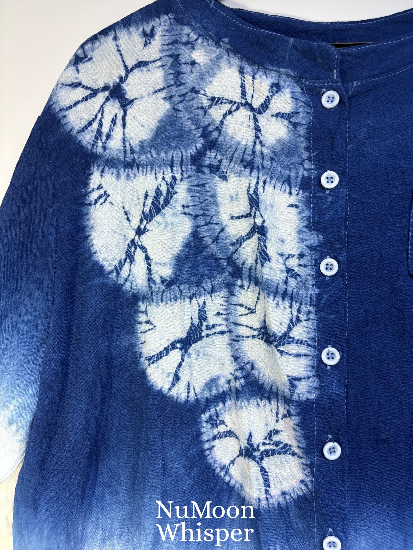 A close-up of a blue and white tie-dye button-up shirt with a collar and white buttons, featuring a circular shibori-style pattern. The brand name 'NuMoon Whisper' is printed in white text at the bottom.