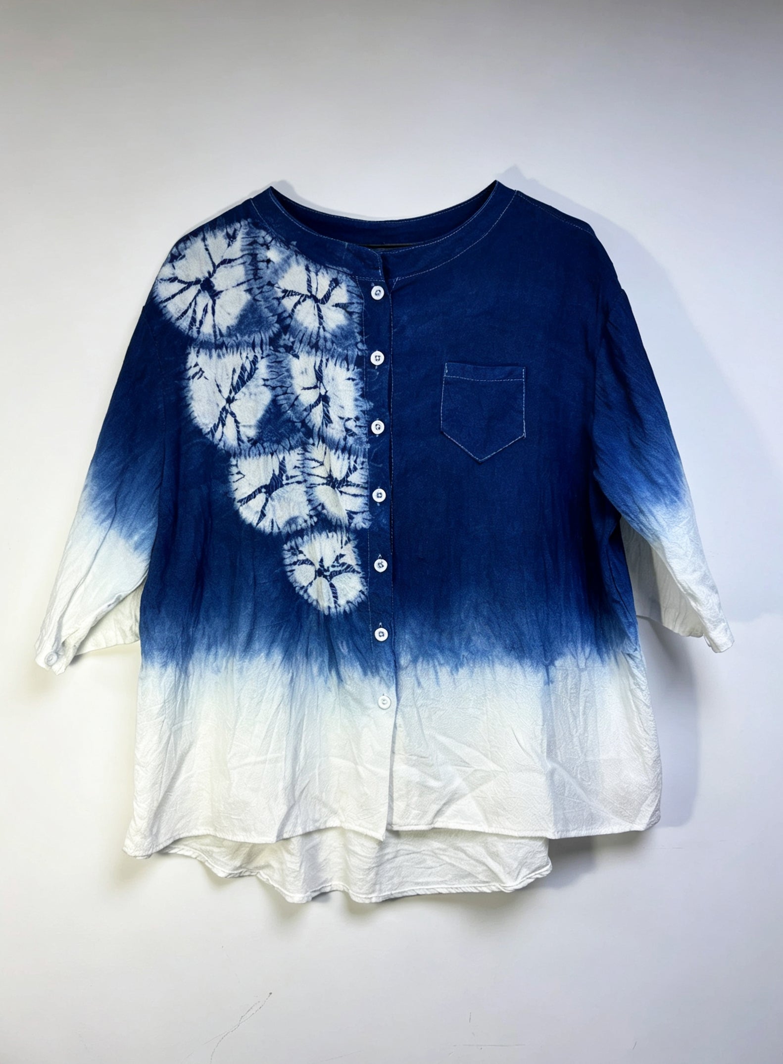 A blue and white tie-dye button-up shirt with a chest pocket, hanging against a plain light background.