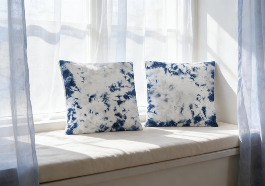 Two blue and white patterned cushions on a light-colored window seat with sheer curtains.