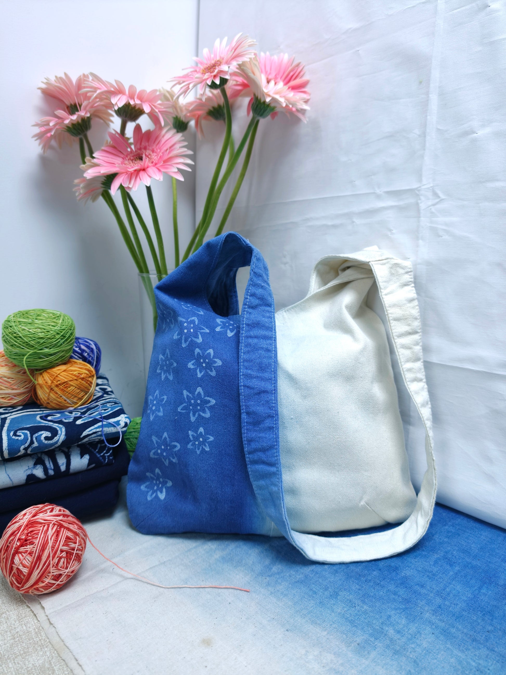 Two fabric bags, one blue and one white, with yarn and flowers on a light blue surface.