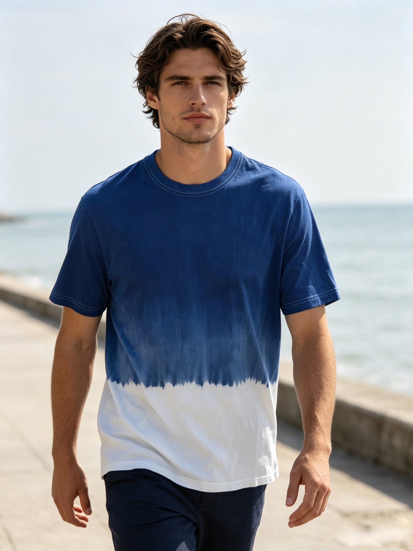 A man wearing a blue and white tie-dye t-shirt walking along a sunny seaside boardwalk.
