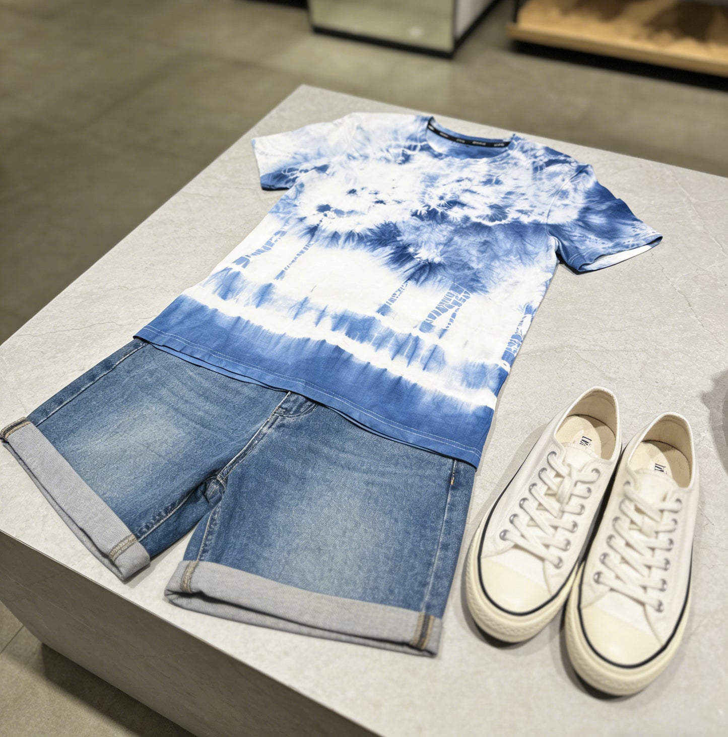 Tie-dye t-shirt, denim shorts, and white sneakers on a light gray surface.