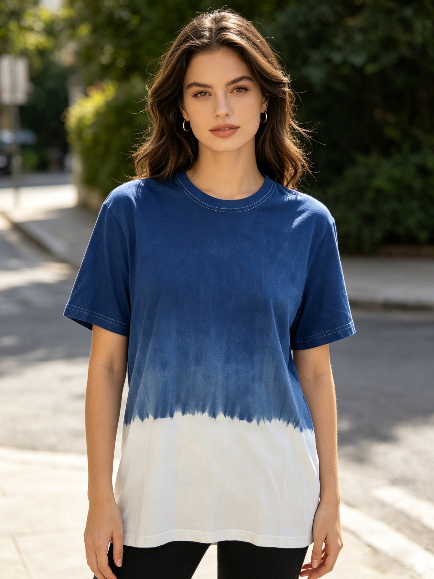 A woman wearing a blue and white tie-dye t-shirt, standing outdoors on a sunny street with blurred green trees in the background.