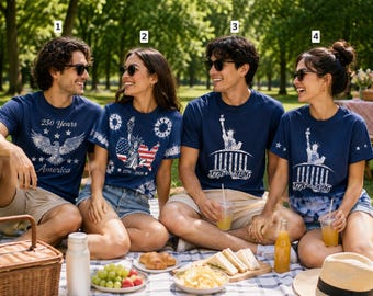 Four people sitting on a picnic blanket outdoors in a park, wearing matching navy blue t-shirts with patriotic US-themed graphics including the Statue of Liberty and American flag motifs.