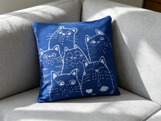 A square blue throw pillow with a white line-art print of multiple cats, resting on a light gray fabric sofa in natural light.